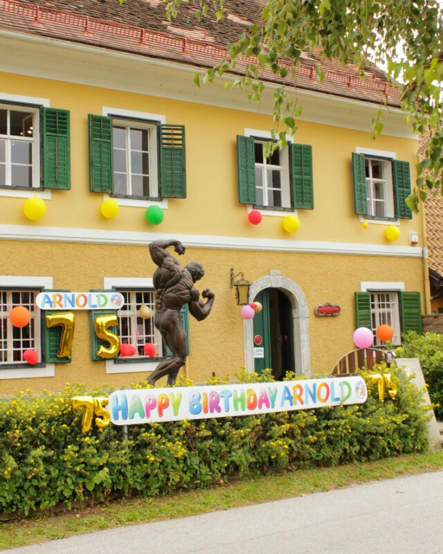 Zum 75. Geburtstag von Arnold Schwarzenegger findet im Schwarzenegger Museum eine Feier statt.  Das Haus und der Garten sind dekoriert. Eine Statue steht am Eingang.